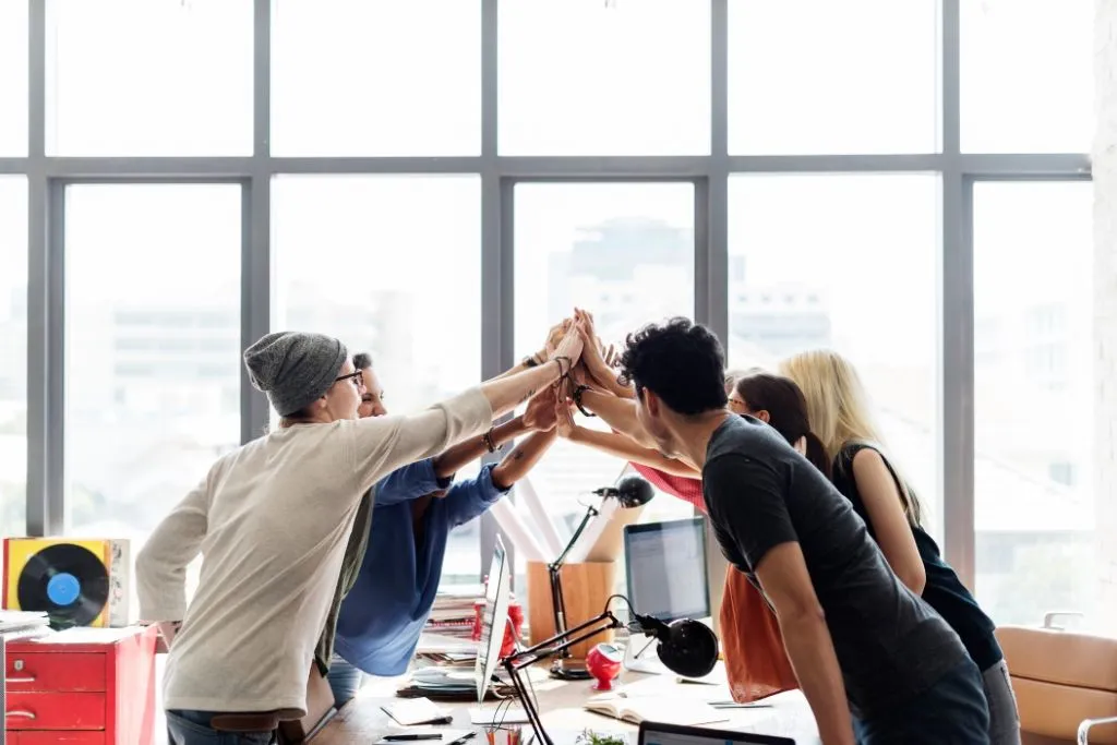 A team of diverse professionals celebrating a successful collaboration with a group high-five in a bright, modern office space, symbolising teamwork and achievement.
