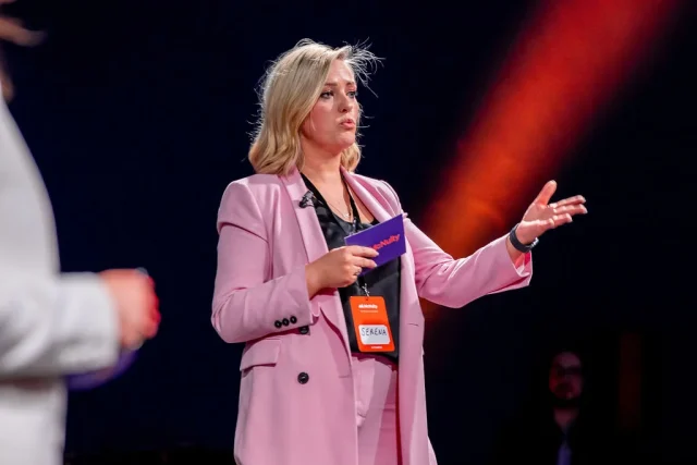 Confident female speaker in a pink blazer presenting at a conference, gesturing with her hands while holding a McNulty business card.