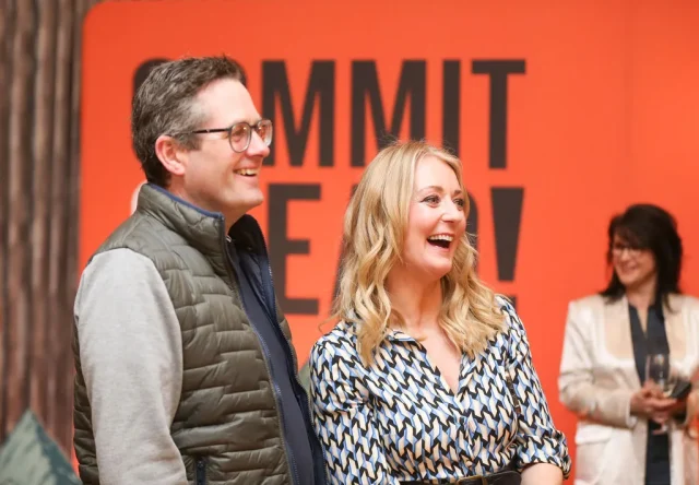 Two people smiling and looking to the side, standing in front of an orange background with the partially visible text 'COMMIT' with another person smiling in the background.