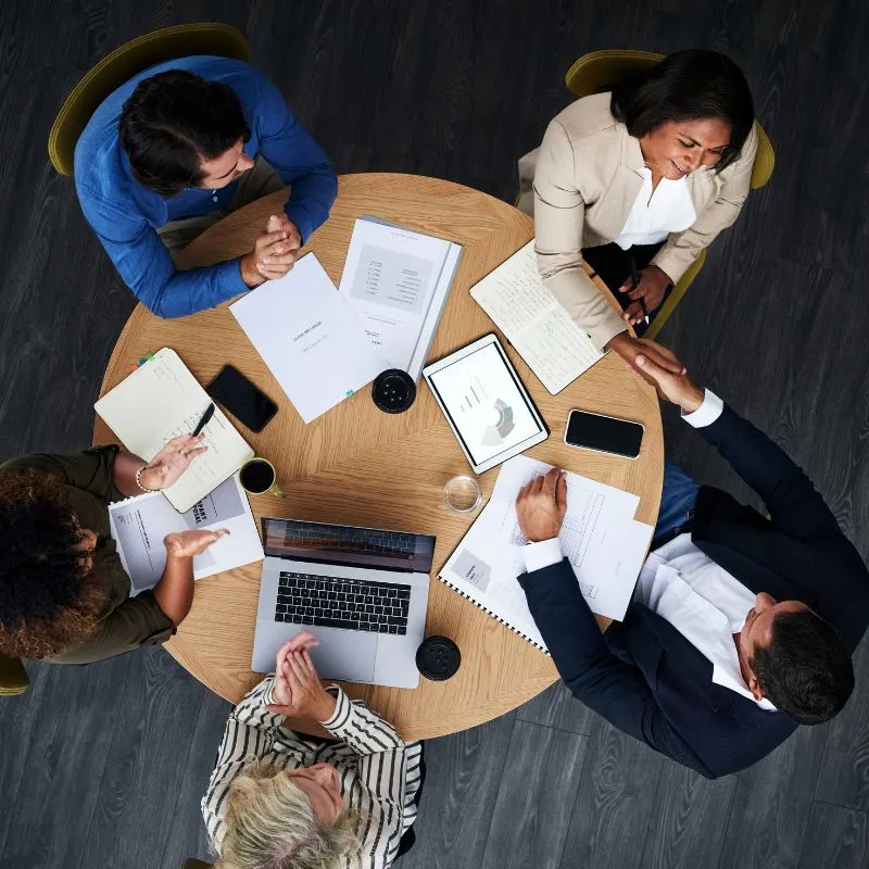 Top-down view of a diverse team engaged in a roundtable discussion with documents, laptops, and smartphones on a wooden table.