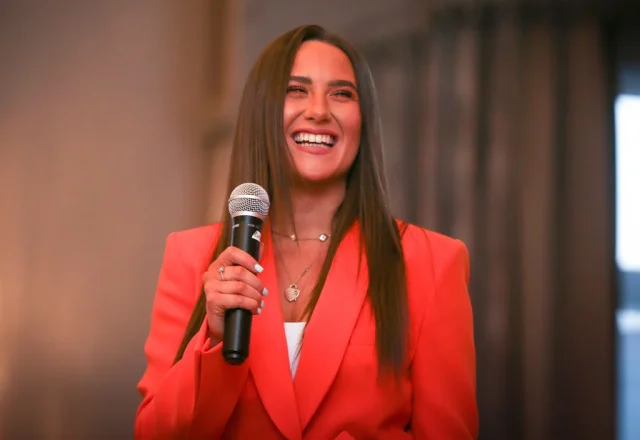 A smiling woman in a bright red suit speaking into a microphone at a professional event, conveying enthusiasm and confidence.
