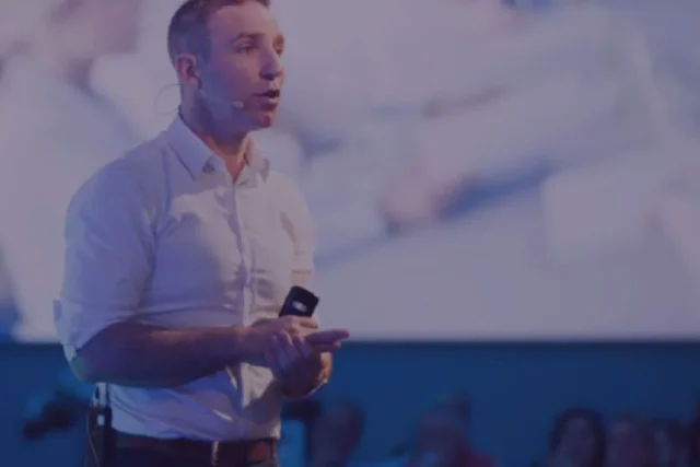 Man presenting at a conference, passionately speaking into a headset microphone, with blurred audience in the background.