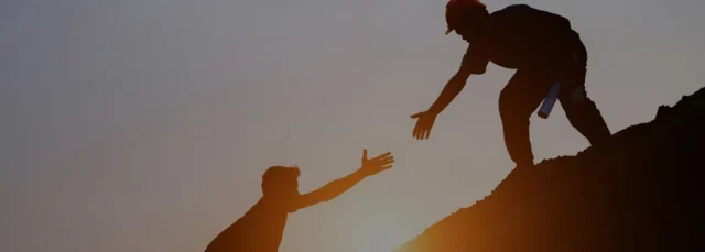 Silhouette of one person reaching down to help another climb up a steep hill during sunset, symbolising support and teamwork in challenging situations.