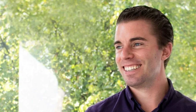 Smiling young man in a casual purple shirt against a blurred green leafy background, radiating positivity and confidence.