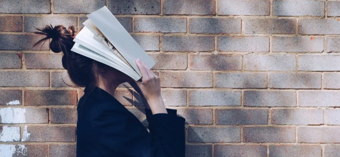A young woman playfully covers her face with books against a brick wall, representing continuous learning and personal development.