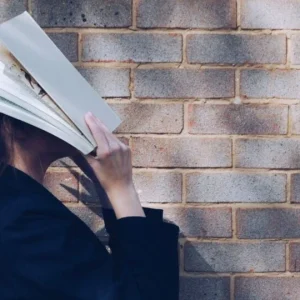 A young woman playfully covers her face with books against a brick wall, representing continuous learning and personal development.