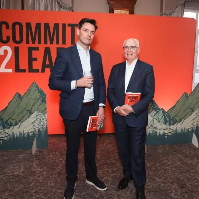 Two professionals with Commit 2 Lead books, standing by event backdrop.