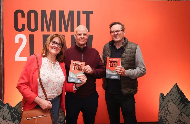 Three participants showcasing their Commit 2 Lead books, posing against an orange backdrop.