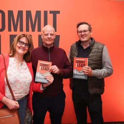 Three participants showcasing their Commit 2 Lead books, posing against an orange backdrop.