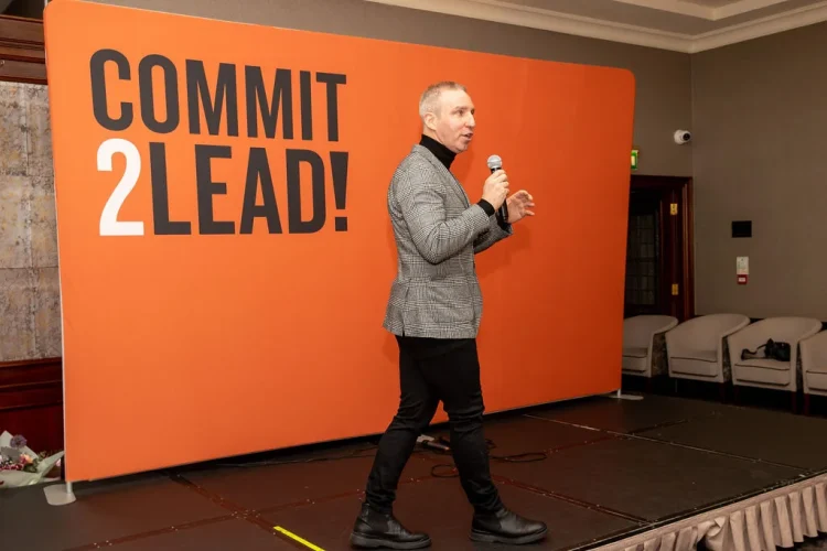 Speaker presenting at Commit 2 Lead event, standing before a large orange backdrop.