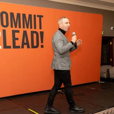 Speaker presenting at Commit 2 Lead event, standing before a large orange backdrop.