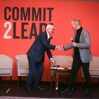 Two men shaking hands at Commit 2 Lead event, one in grey jacket, other in navy suit.