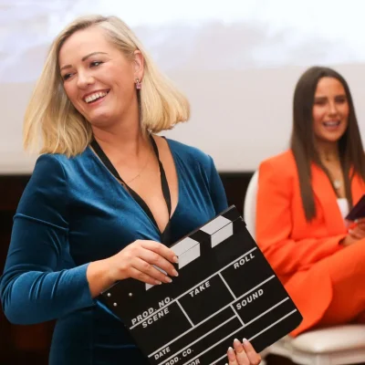 Event coordinator with clapperboard at Commit 2 Lead conference, smiling at audience.