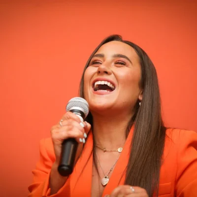 A woman in an orange blazer, smiling and speaking into a microphone at the Commit to Lead event.