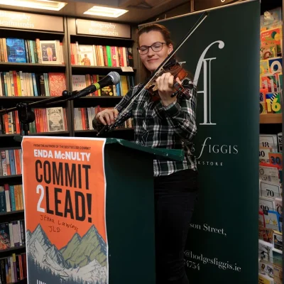 Woman playing violin next to a 'Commit 2 Lead' banner in a bookshop.
