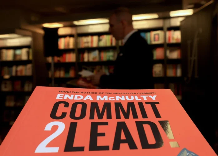 Close-up of 'COMMIT 2 LEAD!' book by Enda McNulty on a desk, blurred shelves in background.