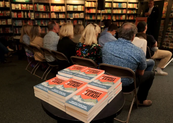 Focused on a stack of 'Commit 2 Lead' books at a Hodges Figgis event with the author speaking in the background.
