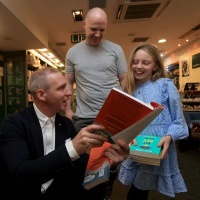 Author showing a book to a young girl, with a man observing at a bookstore event.