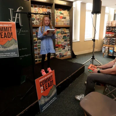 oung girl reading aloud from a book to an audience at a book event.