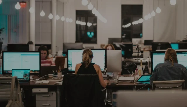 Office scene captured in a lower-light setting, showing various employees engaged at their workstations, highlighting a productive and focused atmosphere.