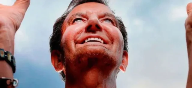 Alan Kelly looking upwards with a smile, raising his hands, against a background of a blue sky.