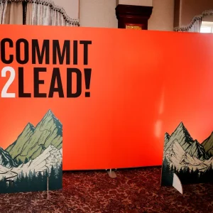 Bright orange banner with the text 'Commit 2 Lead!' in black, flanked by illustrated mountain cutouts, set in an indoor event space.