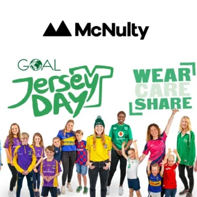 Group of people of various ages wearing different sports jerseys, standing and smiling in front of a banner with the text "Goal Jersey Day" and "Wear Care Share."