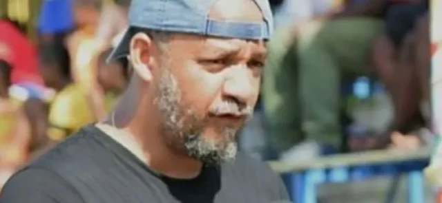 Kako Bourjolly speaking outdoors, wearing a black shirt and a backward baseball cap, with a blurred crowd in the background.