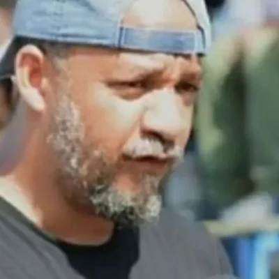 Kako Bourjolly speaking outdoors, wearing a black shirt and a backward baseball cap, with a blurred crowd in the background.