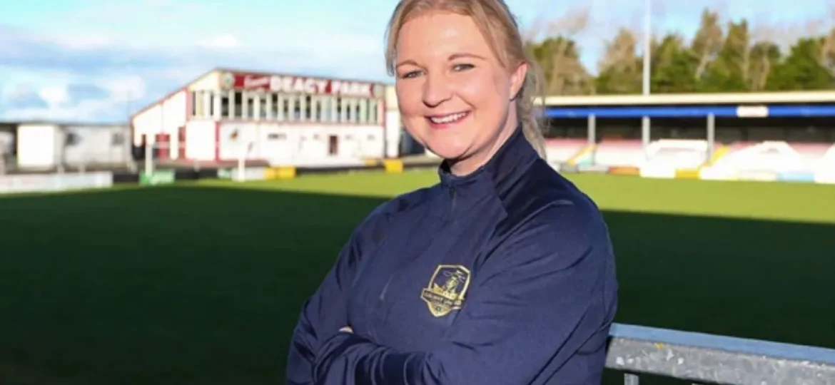 Lisa Fallon standing with her arms crossed on a sports field, smiling, with a stadium in the background.