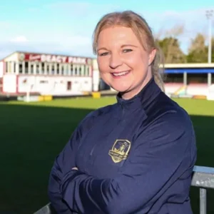 Lisa Fallon standing with her arms crossed on a sports field, smiling, with a stadium in the background.