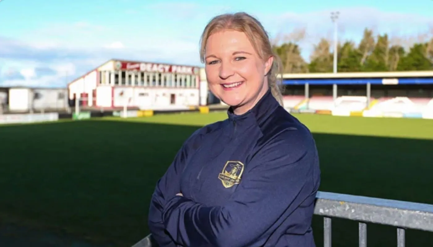 Lisa Fallon standing with her arms crossed on a sports field, smiling, with a stadium in the background.