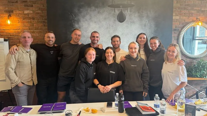 Perpetua Fitness team members and Enda McNulty gathered together after a leadership session, smiling and standing in front of a chalkboard wall with workshop materials on the table in front.
