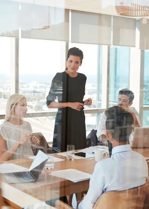 Confident woman standing and addressing a boardroom meeting with colleagues, in a modern office with a scenic city view.